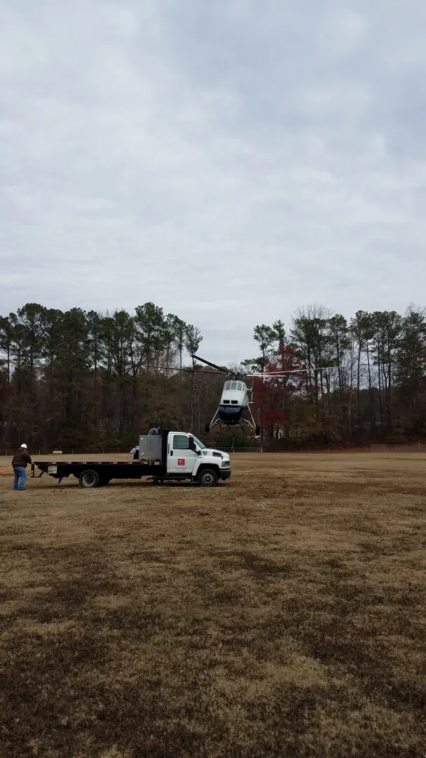 A helicopter lifting off with a truck in the front and trees in the back.